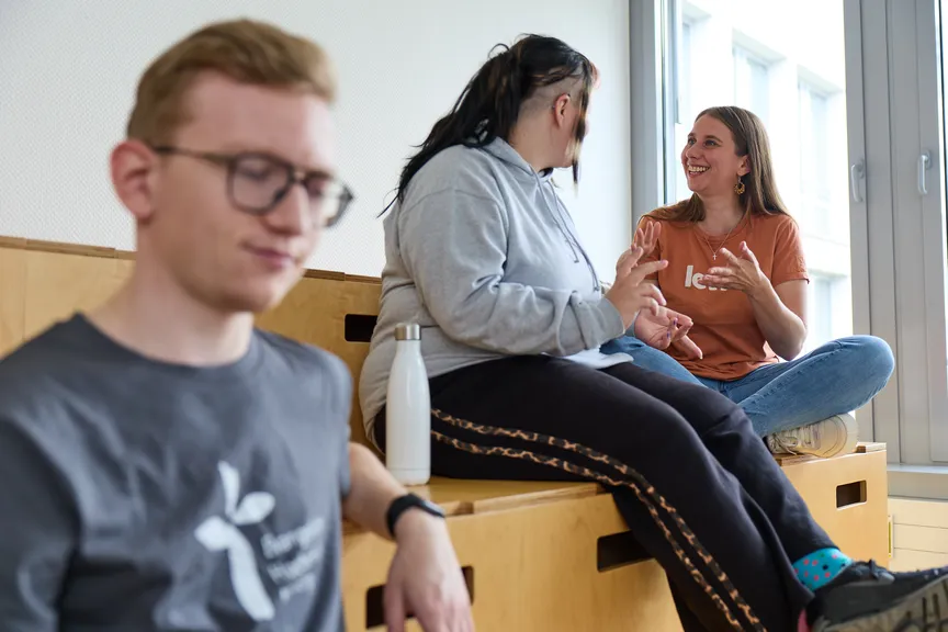 Einige Studierende sitzen auf einer hölzernen Sitzgelegeneheit und unterhalten sich