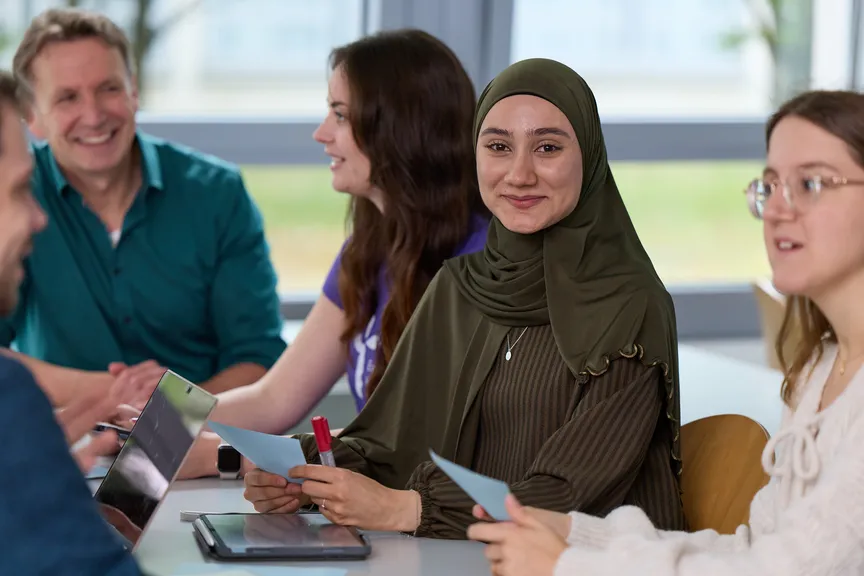 Eine Studierende sitzt in einer Gruppe Studierender in einem Seminar und lächelt in die Kamera